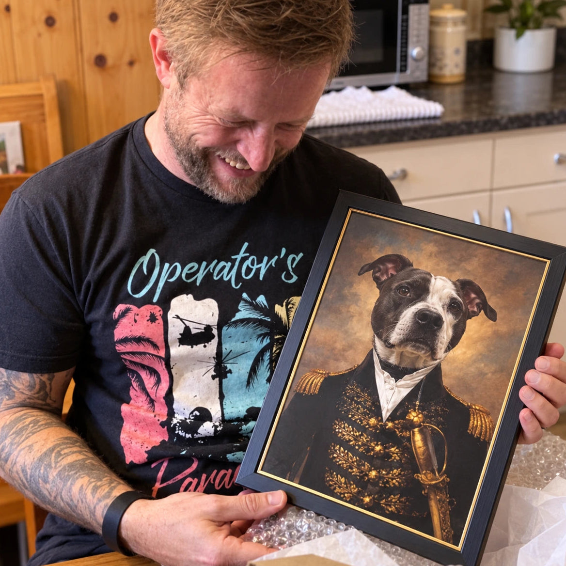 A man in a black graphic t-shirt holds up a framed custom pet portrait of a pitbull-type dog dressed as a military general, with a gold-trimmed black frame and a warm sepia background.
