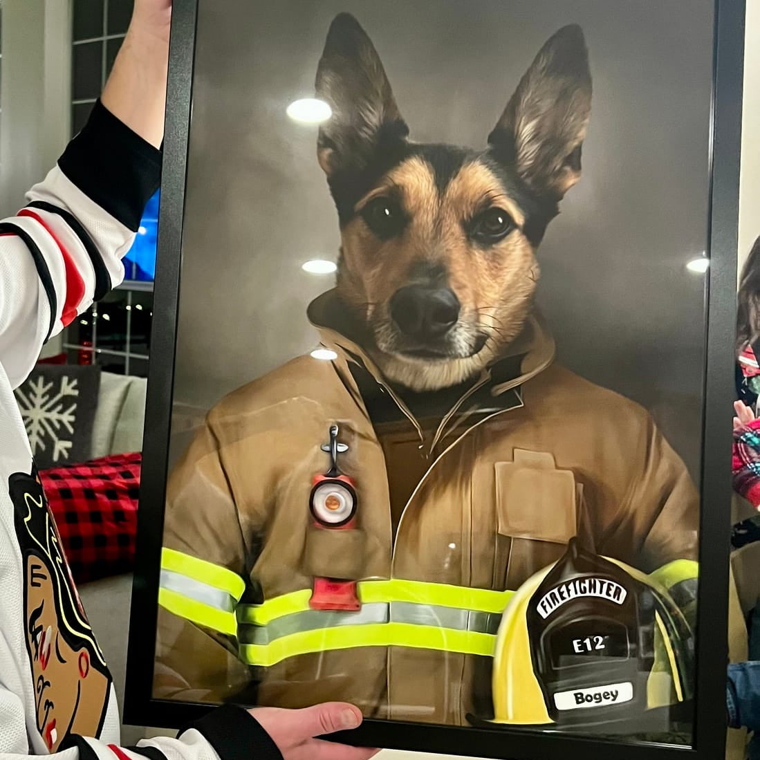 A custom framed pet portrait of a German Shepherd dressed as a firefighter, held up by a person wearing a white and red striped sweater. The portrait features a black frame, a gray background, and a yellow helmet at the bottom right.