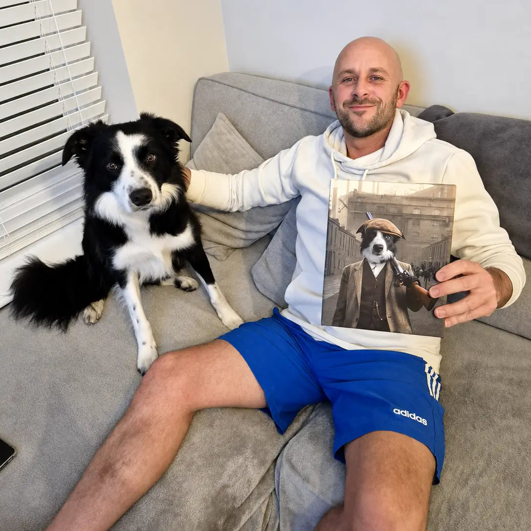 A man sitting on a gray couch with his dog, holding up a custom pet portrait print. The man is wearing a white hoodie and blue Adidas shorts, and the dog is a black and white Border Collie.
