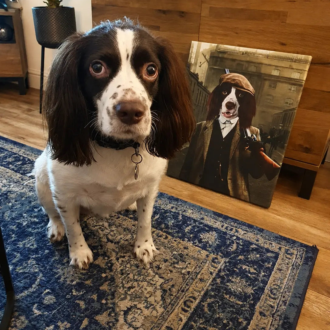A real dog sits on a blue patterned rug in a home interior, looking at the camera. Behind it, a wooden cabinet holds a printed portrait of the same dog dressed in Victorian-era clothing.